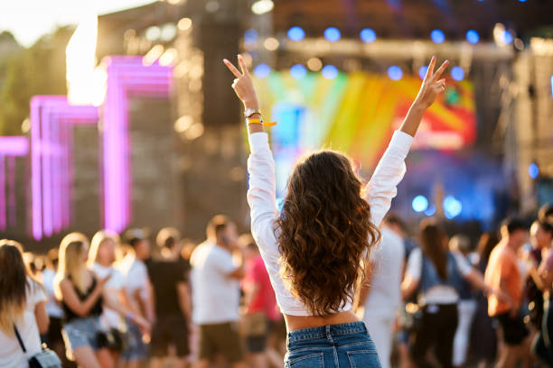 Person at a music festival with raised hands wearing a wristband in front of a colorful stage.